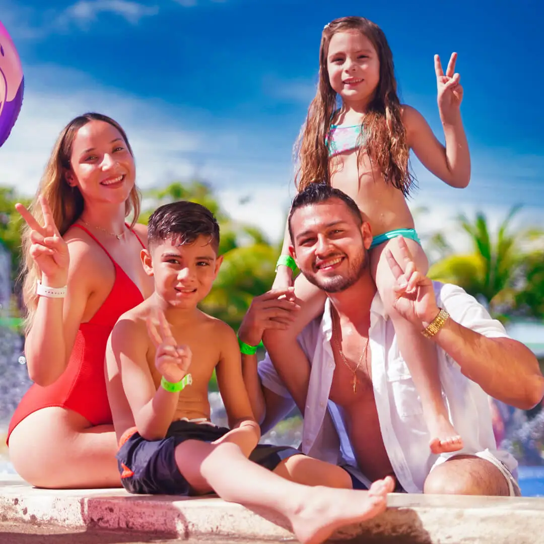 Família composta por mãe, pai e 2 crianças, filho e filha, sorrindo, fora da piscina, acenando com 2 dedos para a foto.